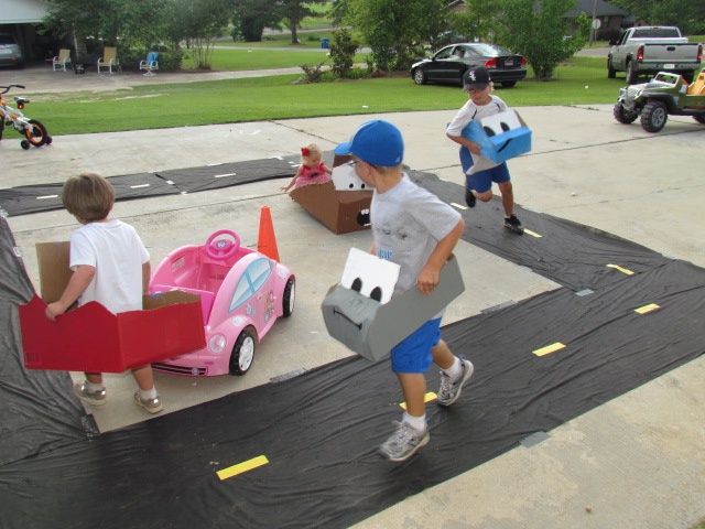 three children are playing with cardboard cars in the driveway