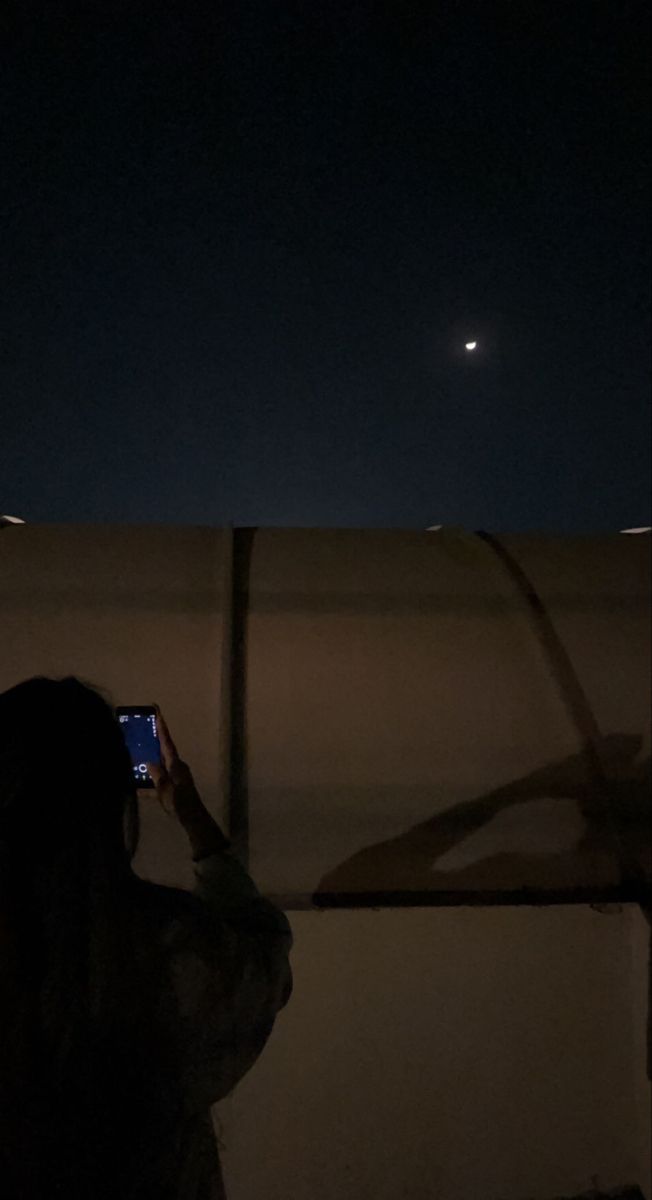 a woman taking a photo with her cell phone at night, while the moon is in the distance