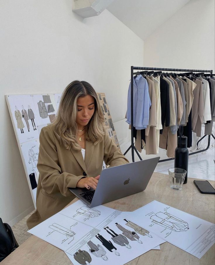 a woman sitting at a table with a laptop computer in front of her, surrounded by clothes