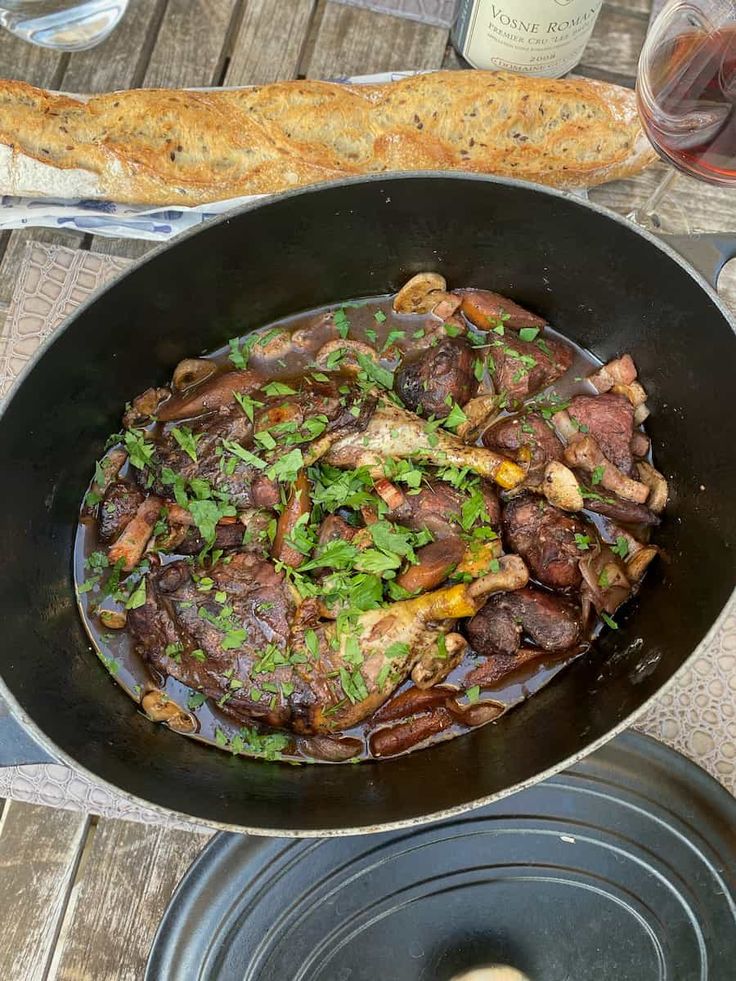 a pan filled with meat and vegetables on top of a wooden table next to bread