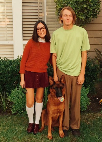 a man and woman standing next to a dog in front of a house with bushes