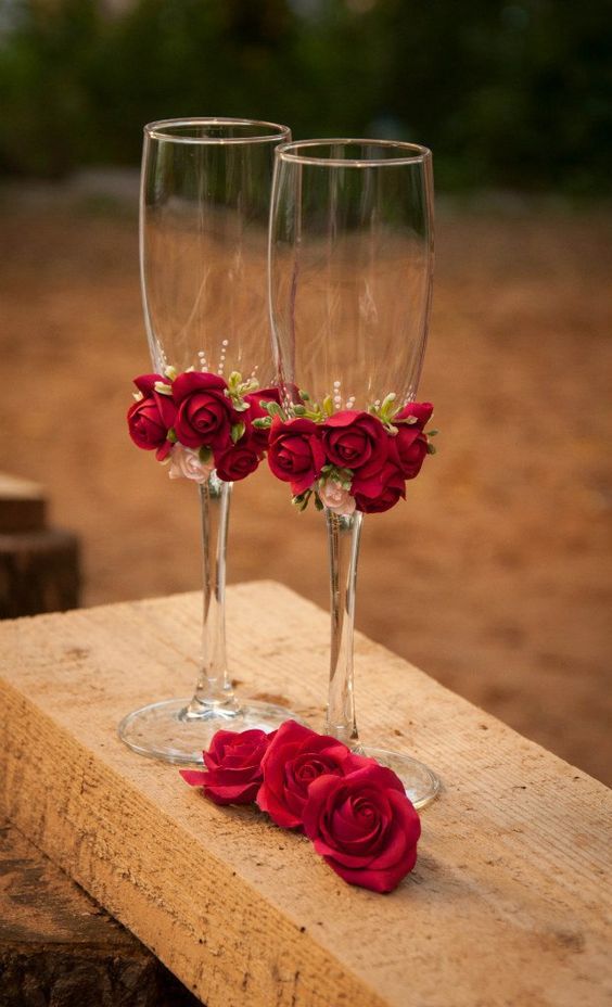 two wine glasses with red roses in them are sitting on a piece of wooden plank