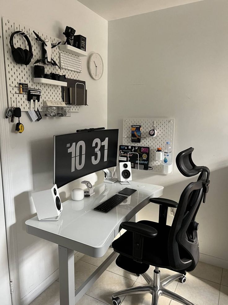 a white desk topped with a computer monitor next to a black chair and clock on the wall