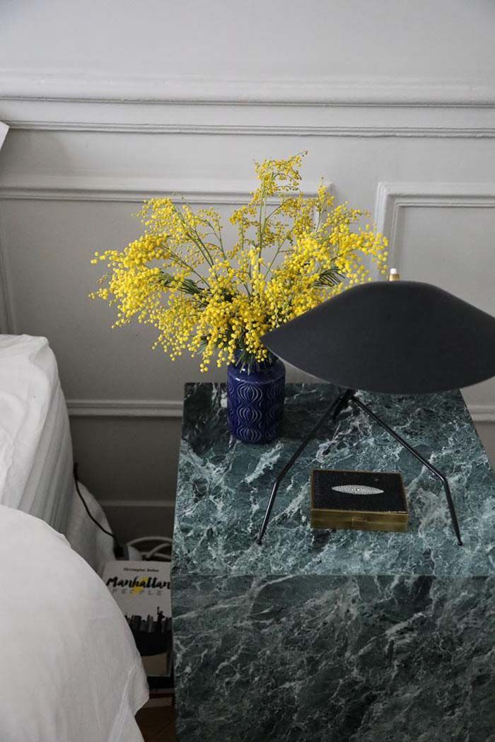 a vase with yellow flowers sitting on top of a green marble table next to a bed