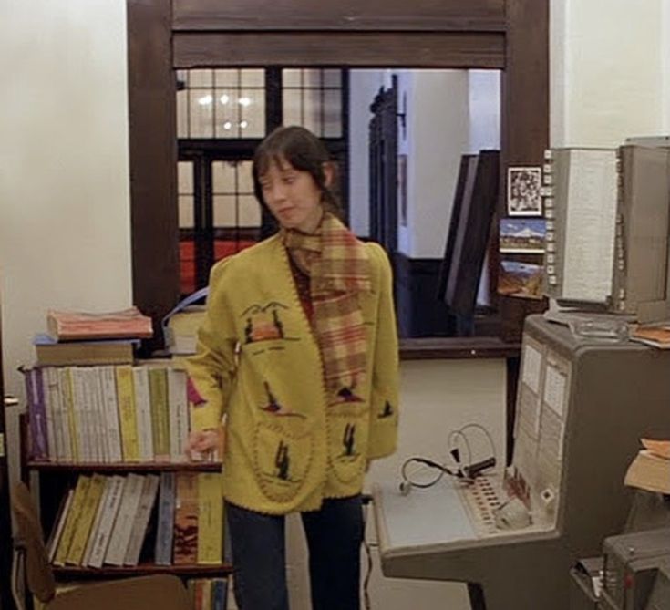 a woman standing in front of a computer desk with books on top of it and an old typewriter behind her