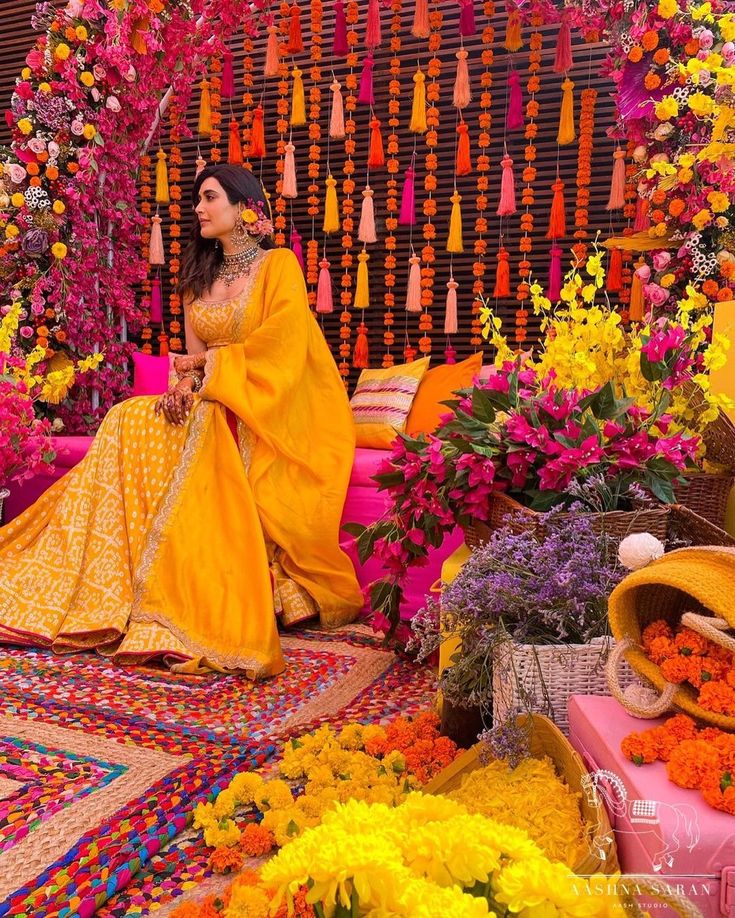 a woman in yellow sitting on a colorful carpet with flowers and decorations around her,