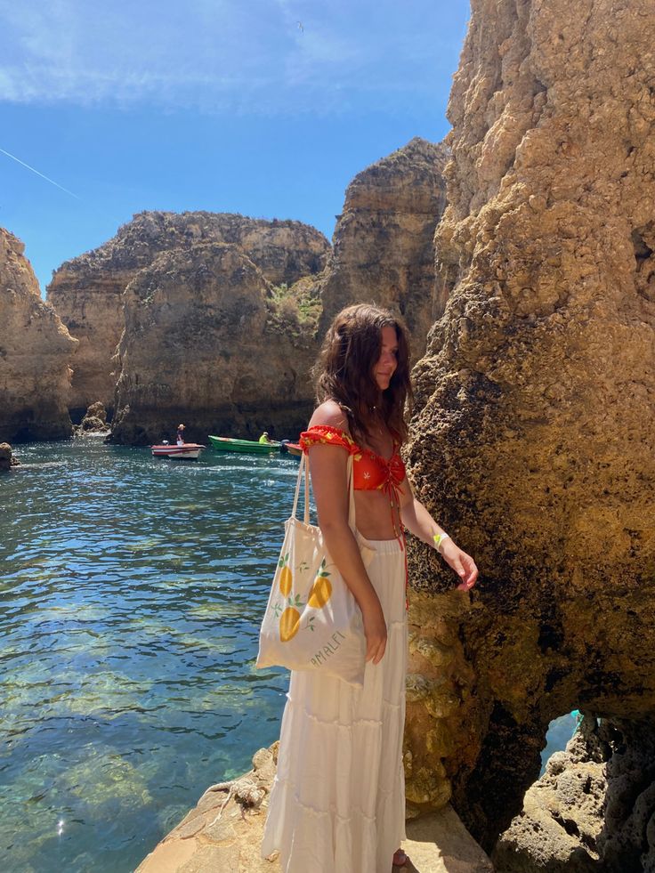 a woman in a white dress is standing on the rocks near the water and holding a bag