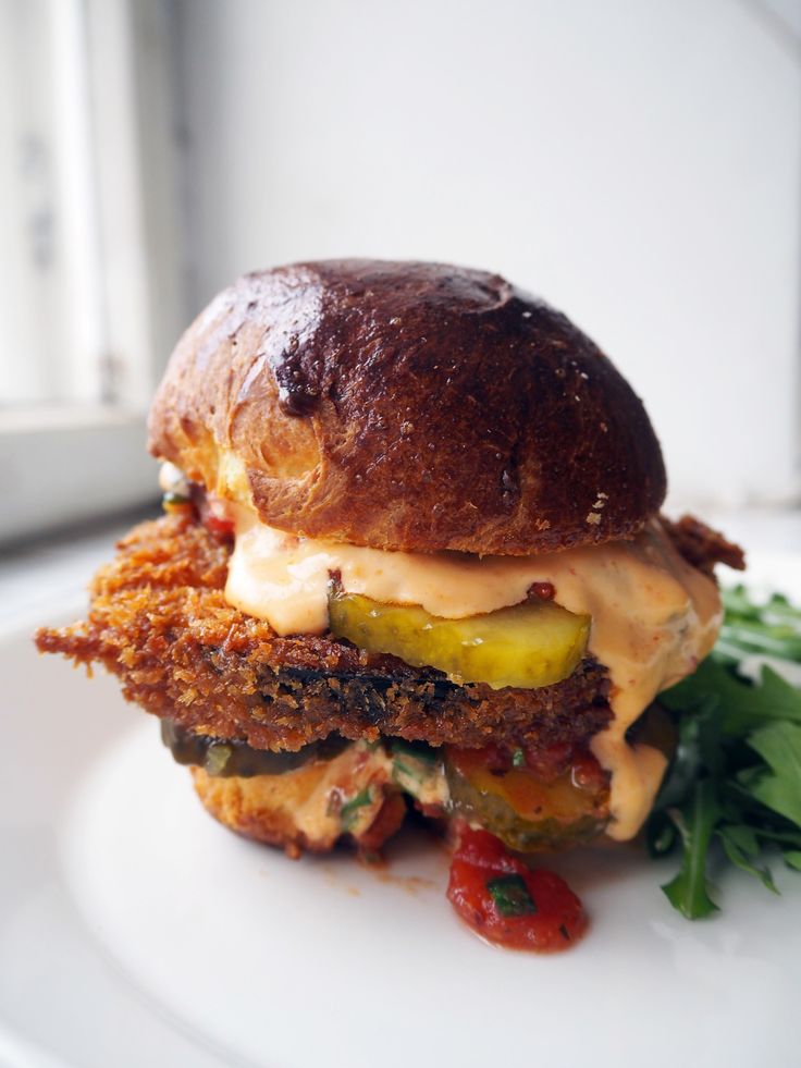 a sandwich with meat, cheese and vegetables on a white plate next to a window