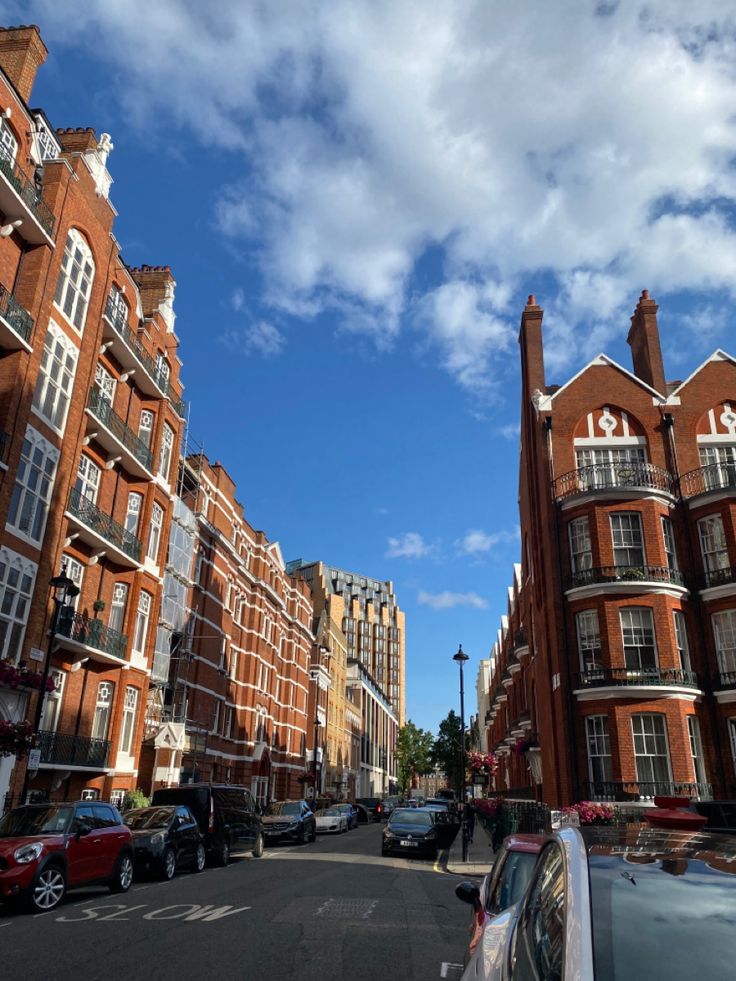 cars parked on the side of a street next to tall red brick buildings under a blue sky with white clouds