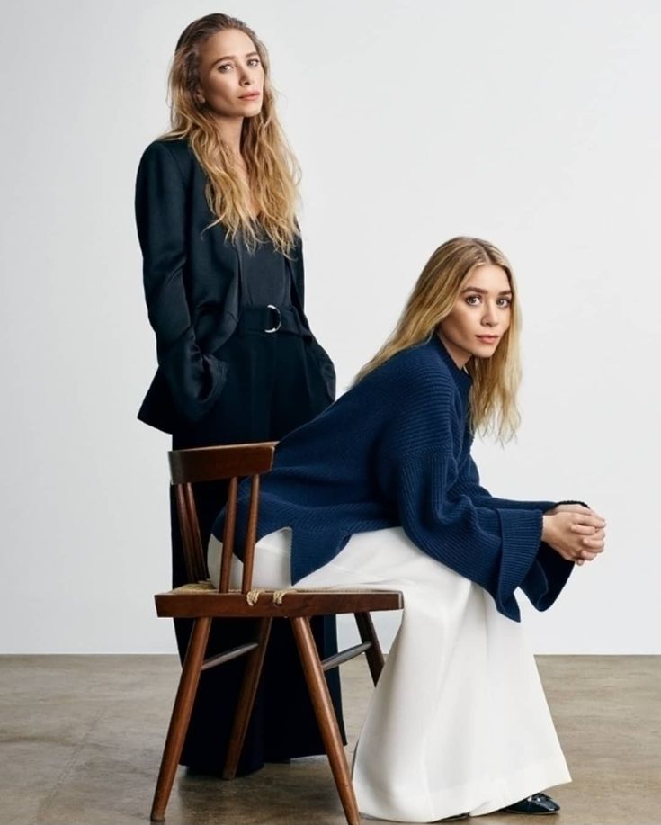 two women are sitting on chairs in front of a white wall and one is wearing a black coat