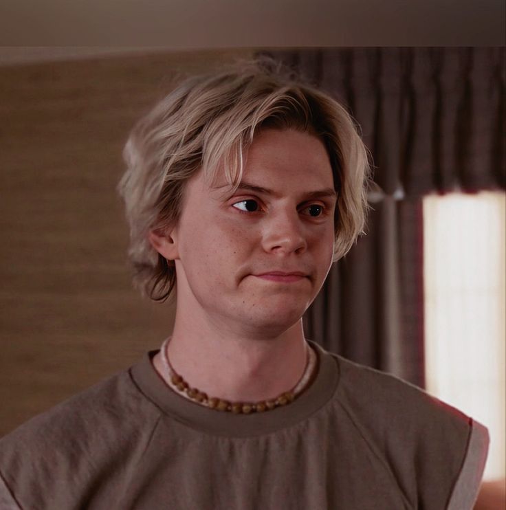 a man with blonde hair wearing a necklace and looking at the camera while standing in front of a window