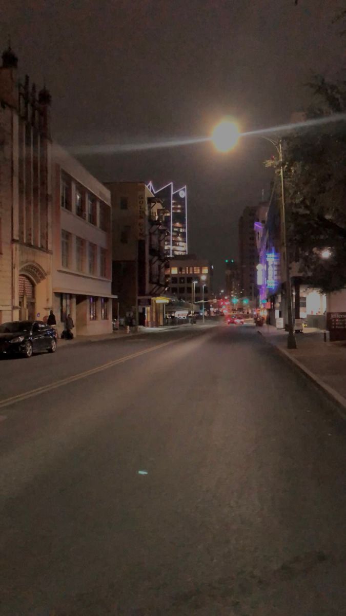 an empty city street at night with cars parked on the side and buildings in the background