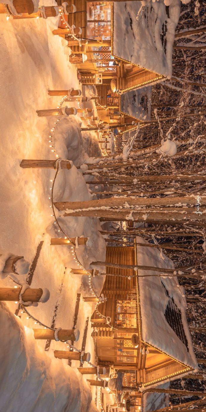 a row of wooden buildings with lights on them in the middle of snow covered ground