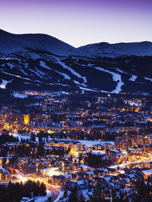 the city is lit up at night with snow on the mountains in the foreground