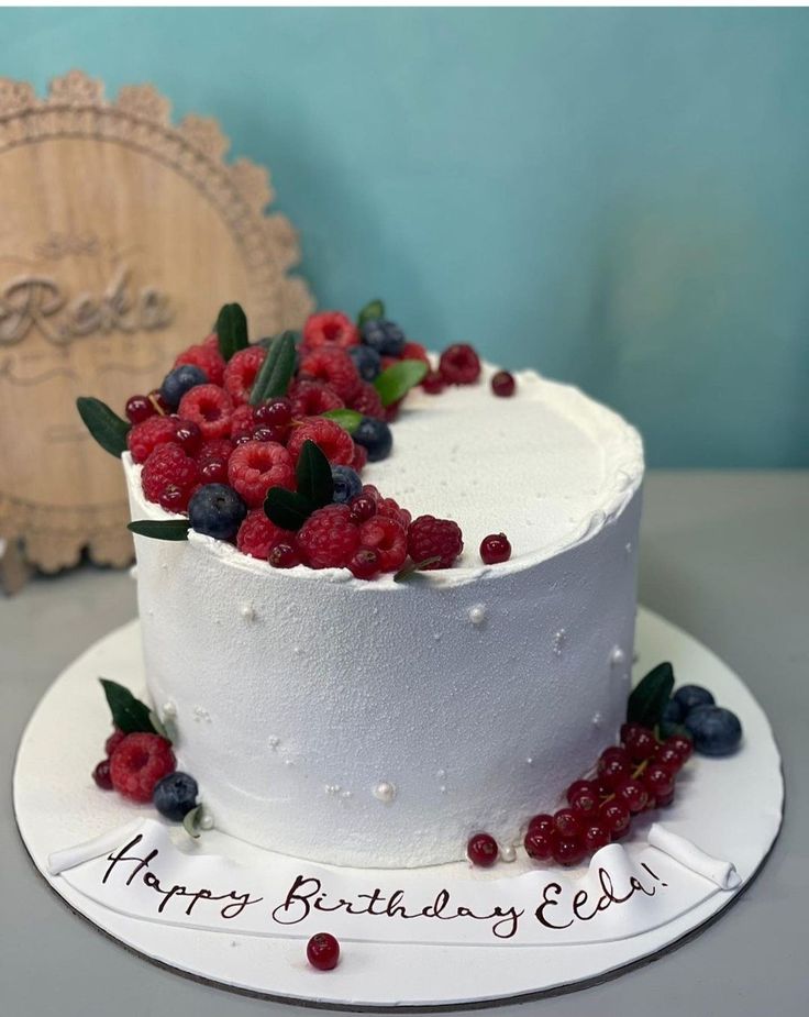 a white cake with berries and leaves on top sitting on a table next to a wooden sign