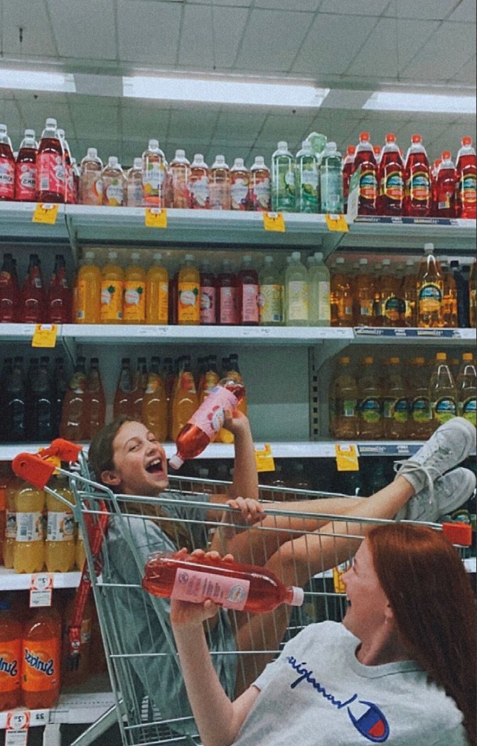 a man and woman sitting in a shopping cart at a grocery store with bottles of soda behind them