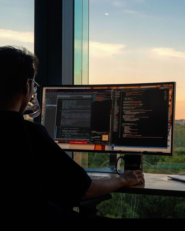 a man sitting at a desk with two computer monitors in front of him and the window behind him