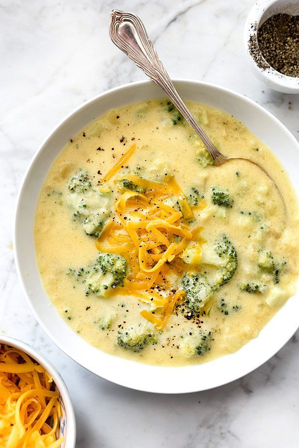 a white bowl filled with broccoli cheese soup next to two bowls of noodles