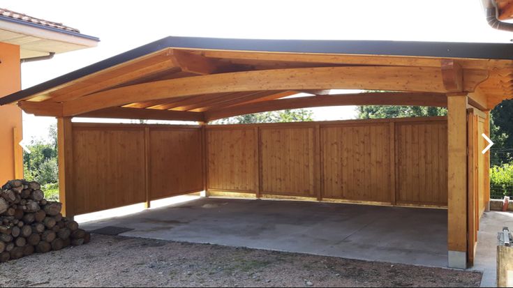 a wooden carport in front of a house with wood logs stacked on the ground