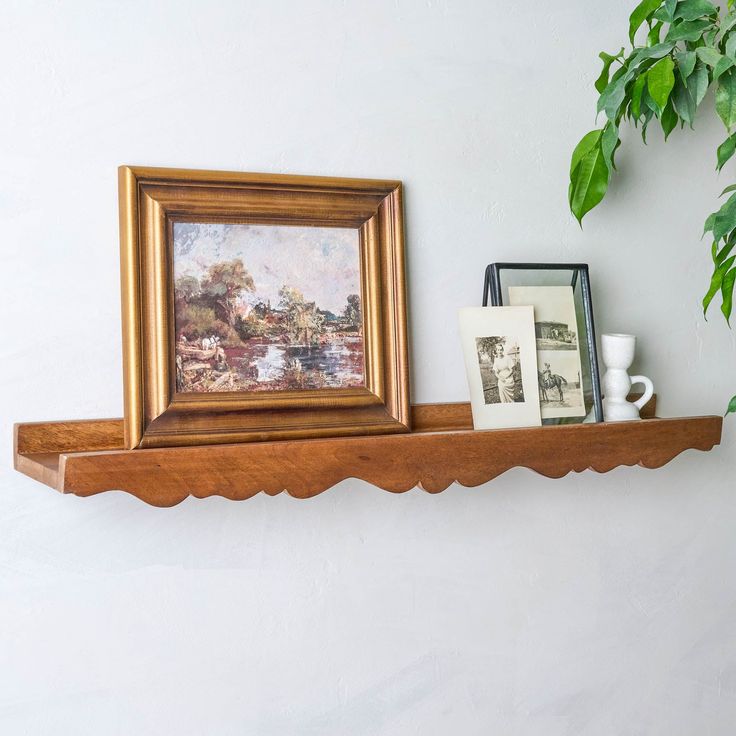 a shelf with two pictures on it and a framed painting hanging above the shelf, next to a potted plant