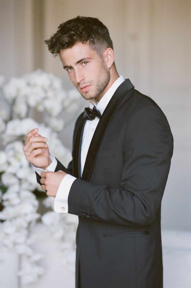 a man in a tuxedo standing next to a flower filled vase with white flowers