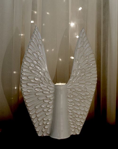 a white vase with an angel wings decoration on it's side next to curtains