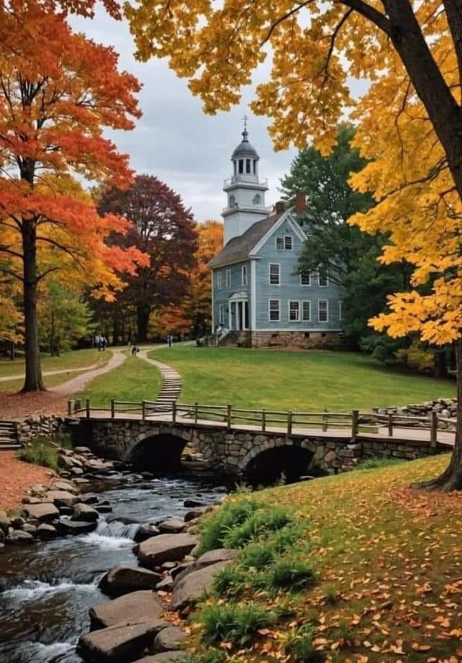 a small bridge crosses over a stream in front of a white house with a steeple on top