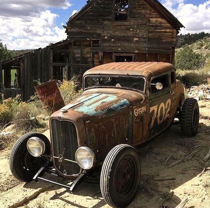 an old rusted out car sitting in front of a wooden building on the side of a dirt road