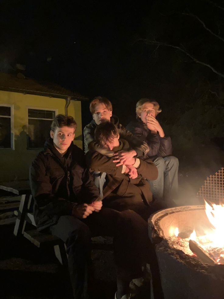 four people sitting around a fire pit at night with one person holding his head in his hands