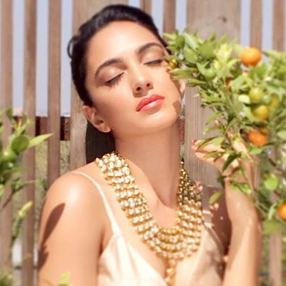 a woman in a white dress holding an orange tree with her eyes closed while wearing a gold necklace