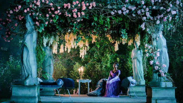 a woman in a purple dress sitting on a bench under an arbor with flowers and candles