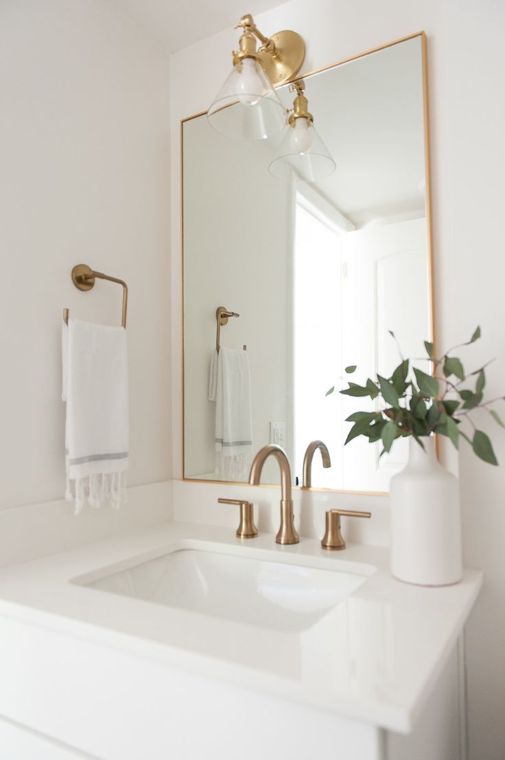 a white bathroom with gold fixtures and a large mirror above the sink that has a plant in it