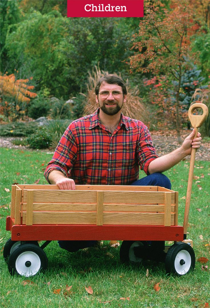 a man sitting in the grass with a wooden wagon holding a rake and wearing a plaid shirt