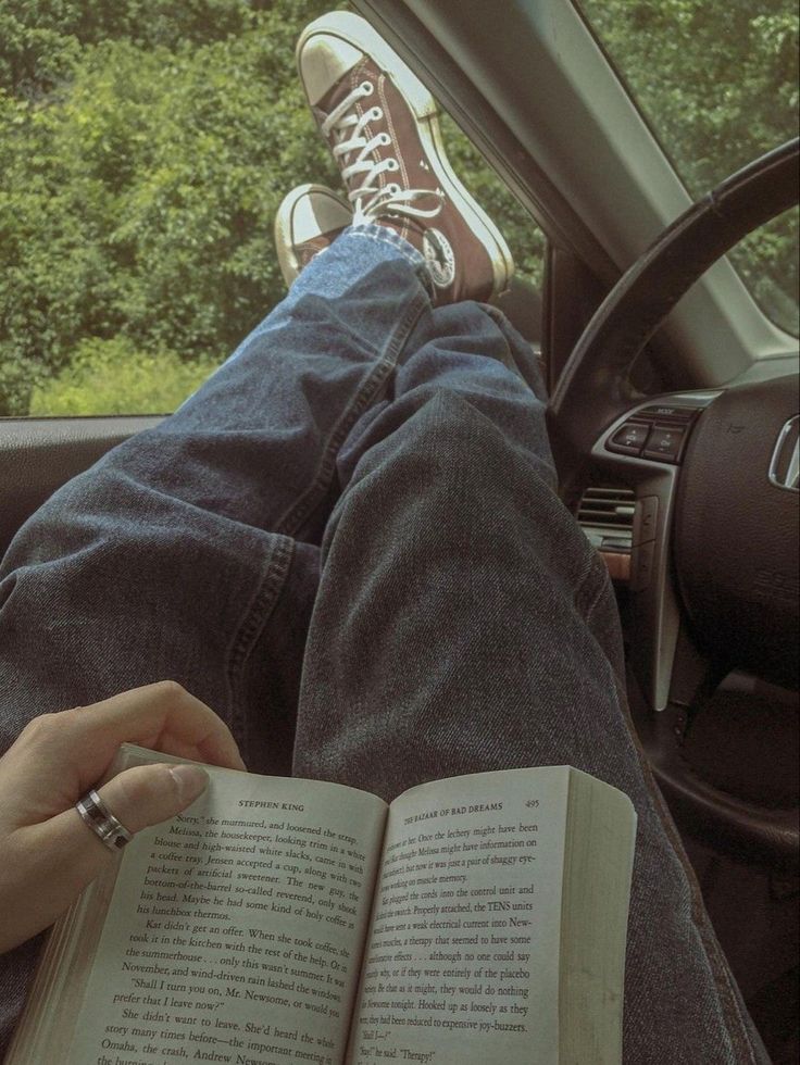 a person sitting in a car with their feet on the steering wheel while reading a book