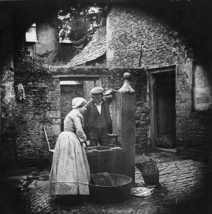 an old black and white photo of two women filling water from buckets in front of a brick building