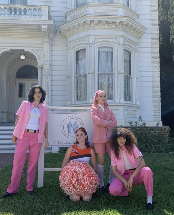 three women in pink outfits posing for a photo with a white house behind them and one woman sitting on the ground