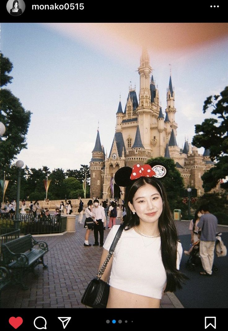 a woman standing in front of a castle with minnie mouse ears on her head and people walking around