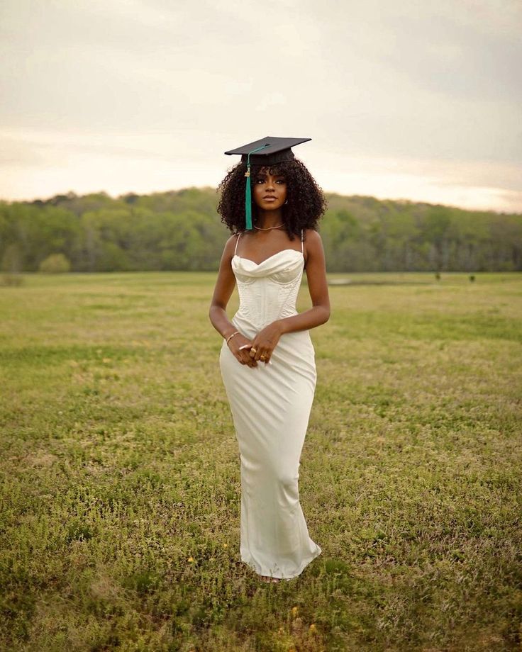 a woman in a white dress and black graduation hat standing in a field with her hands on her hips