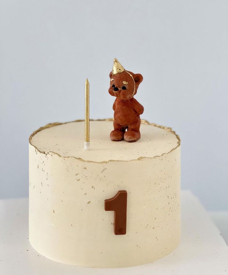 a small teddy bear on top of a white cake with a candle in the shape of a 1