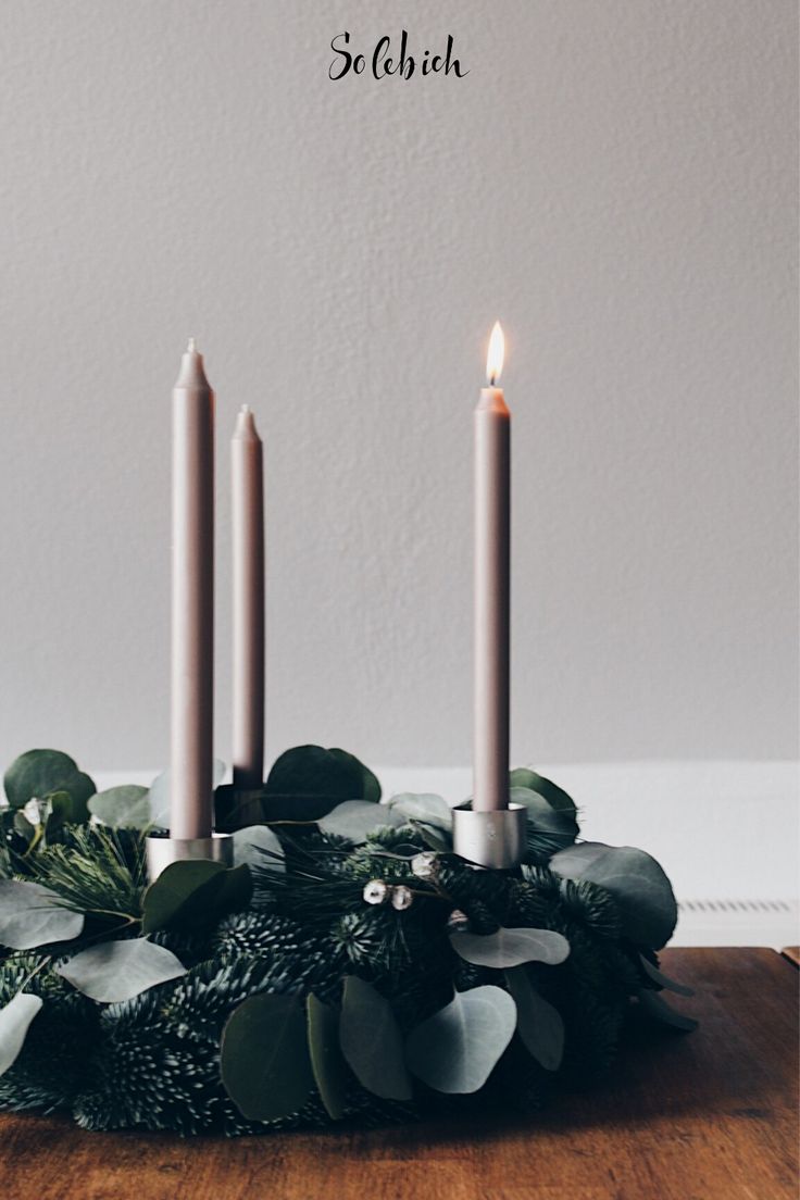 three candles sitting on top of a wooden table with greenery and leaves around it