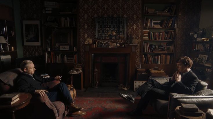 two men sitting on couches in a living room with bookshelves and fireplace