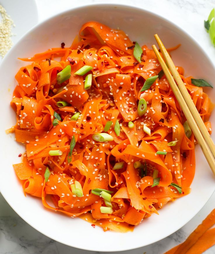 a white bowl filled with carrot noodles and chopsticks next to some sesame seeds