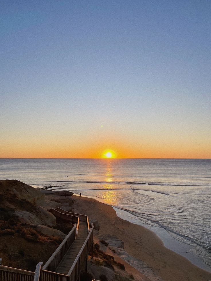 the sun is setting over the ocean with stairs going up to it and people walking on the beach