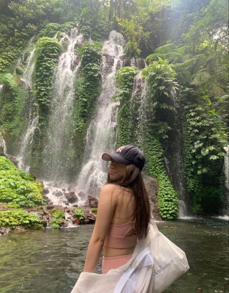 a woman standing in front of a waterfall with a bag over her shoulder and wearing a hat