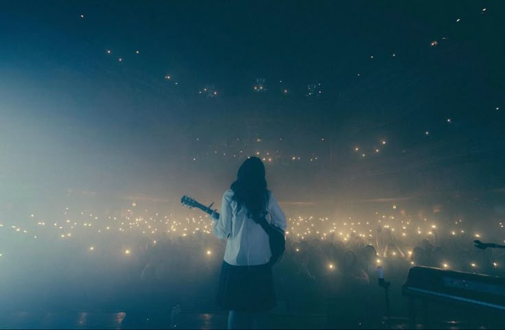 a woman standing in front of a stage holding a guitar and looking at the crowd