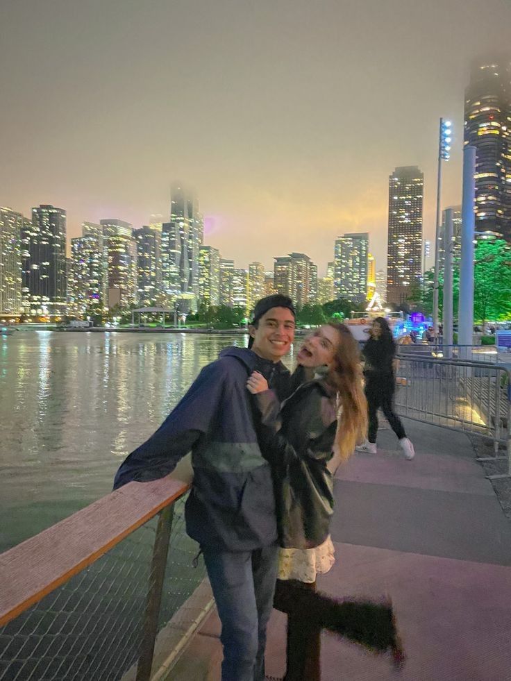 a man and woman standing next to each other on a bridge over water with city lights in the background