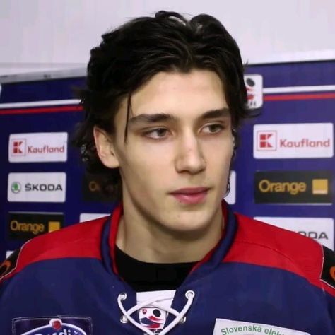 a young man in a blue and red jersey is talking to the camera while standing next to a wall