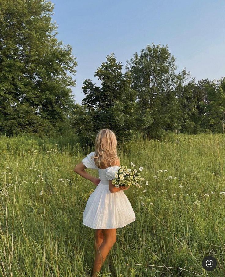 a woman in a white dress is standing in tall grass and holding a bouquet of flowers