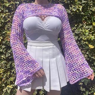 a woman standing in front of a bush wearing a purple crochet sweater and white skirt