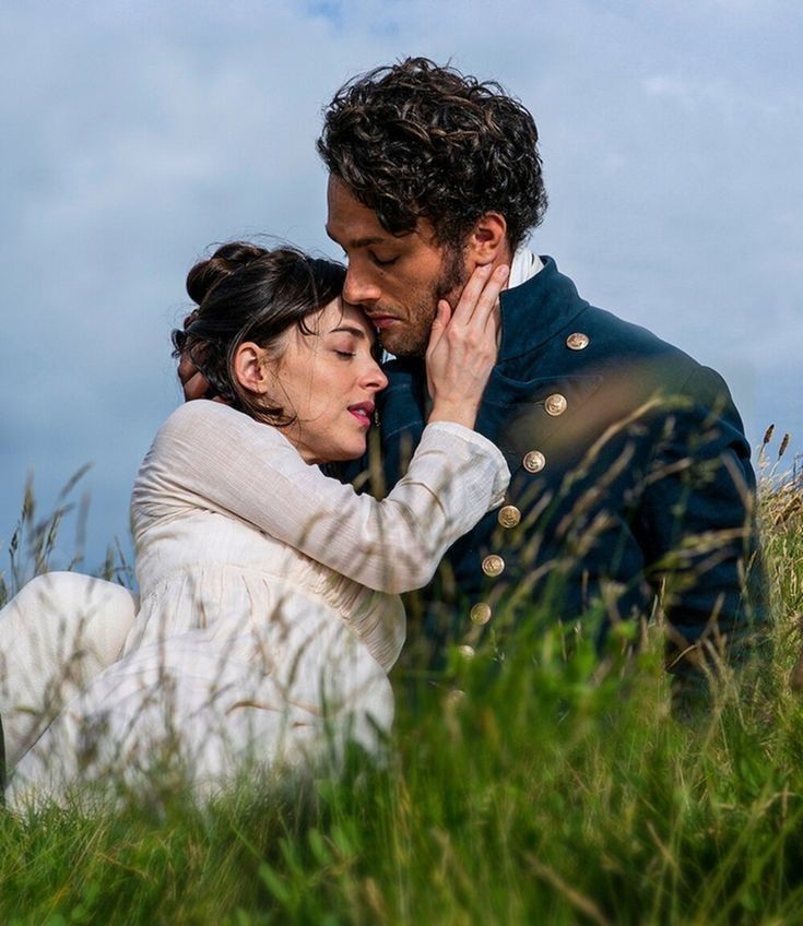 a man and woman standing next to each other in tall grass with the sky behind them